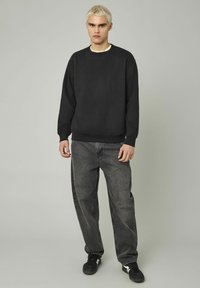 Sweatshirt noir à col rond, coupe ample et poignets côtelés ; associé à un jean gris à coupe décontractée et des baskets noires.