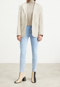 Blazer de lino beige, cuello alto beige, jeans ajustados de color azul claro y botines de tacón bloque con paneles en contraste beige y negro.
