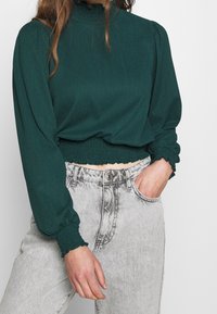 Teal texturerad blus med hög hals, puffärmar och smockdetaljer, bärs med grå högmidjade jeans.