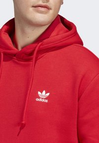 Röd fleece-hoodie med snoddhuva, prydd med en vit Adidas-logotyp på bröstet. Slät textur och avslappnad passform.