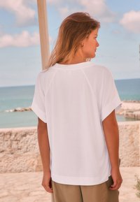 Chemise blanche à manches courtes en tissu léger, avec une coupe décontractée et des fentes latérales, portée avec un pantalon beige sur un fond côtier.