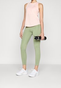 Haut sans manches rose clair, leggings verts avec des détails de couture et chaussures de sport blanches. Bouteille d'eau noire à la main.
