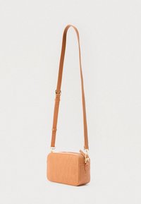 Liten tverrveske i lysebrunt skinn med preget bokstavtrykk og justerbar stropp, vist hengende mot en ensfarget hvit bakgrunn.