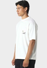 Valkoinen puuvillapaita lyhyillä hihoilla ja rintataskulla, jossa on värikäs graafinen kuvasto. Sisältää pienen lapun hihassa.