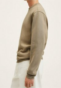 Persona che indossa una felpa beige a maniche lunghe e pantaloni bianchi, in piedi di lato contro uno sfondo chiaro e uniforme.