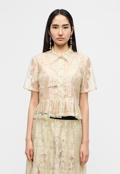 Μπλούζα με κοντά μανίκια από διάφανο δαντελένιο ύφασμα σε ανοιχτό μπεζ χρώμα με λουλουδένια σχέδια, κλείσιμο με κουμπιά και τελείωμα με πεπλούμ. Φορεμένη πάνω από φούστα που ταιριάζει.