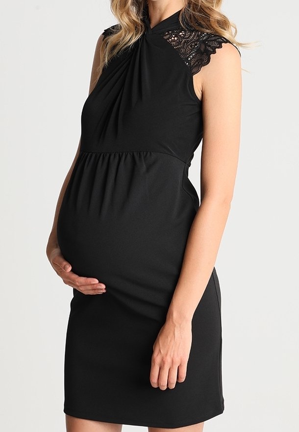 Femme enceinte en robe noire sans manches avec un détail en dentelle sur l'épaule, tenant son ventre arrondi d'une main, debout devant un fond uni.