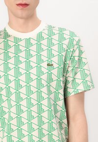 Tricou alb cu mânecă scurtă, cu un model geometric verde, având un mic logo verde de crocodil în partea stângă. Fabricat dintr-un material ușor.