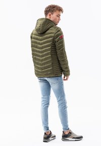 Giacca a vento verde oliva con cappuccio, caratterizzata da trapuntatura orizzontale e verticale, con una patch rossa sul manico, abbinata a jeans chiari e sneakers.