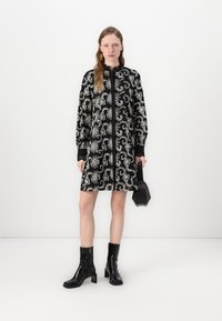Veste preta com um intrincado padrão de espirais brancas, apresentando mangas longas e um fecho na frente. Combinada com botas de tornozelo pretas e uma pequena bolsa.