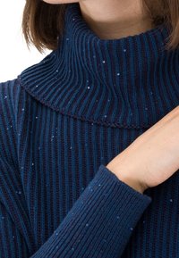 Mujer llevando un suéter de lana de cuello alto de color azul marino con pequeñas lentejuelas, mostrando parte de su cuello y un brazo cruzado sobre su pecho.