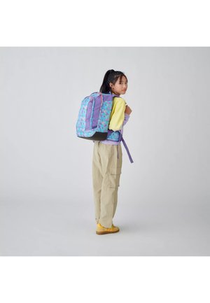 Jeune femme portant un sac à dos géométrique coloré, des chaussures jaunes, un pantalon beige et un sweat-shirt pastel violet et jaune, debout de profil.