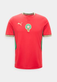 Κόκκινη φανέλα ποδοσφαίρου με κοντά μανίκια και πράσινες λεπτομέρειες, λογότυπο Puma και έμβλημα της Μαροκινού ποδοσφαιρικής ομοσπονδίας στο στήθος.