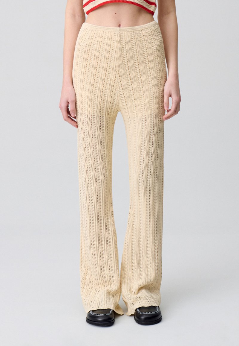 Pantalones de pierna ancha de color crema, tejidos, que presentan una textura de canalé vertical y una cintura alta, combinados con zapatos planos negros.