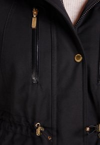 Schwarze Jacke mit goldfarbenen Beschlägen, Reißverschlusstaschen und tailliertem Schnitt. Glatte Textur mit klarer Linienführung und strapazierfähigem Stoff.