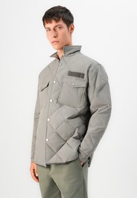 Gray quilted jacket with a high collar, snap buttons, and two chest pockets. Features a textured fabric and a branded patch on the front.