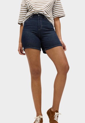 Person trägt dunkelblaue hoch taillierte Jeansshorts, ein gestreiftes beige-weißes kurzärmeliges Hemd und braune Schnürschuhe, steht vor einem einfarbigen Hintergrund.