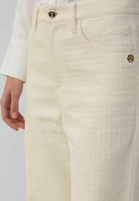 Pantalon en denim couleur crème avec un motif texturé, poches avant, un bouton doré et un discret emblème de marque sur la ceinture.