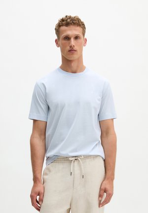 Jonge man met krullend haar, gekleed in een lichtblauw T-shirt met korte mouwen en beige broek met trekkoorden, tegen een effen witte achtergrond.