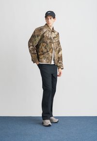 Bunda s maskáčovým vzorom v hnedej a zelenej farbe, kombinovaná s bielym tričkom, čiernymi nohavicami a sivými teniskami. Model nosí čiernu čiapku.