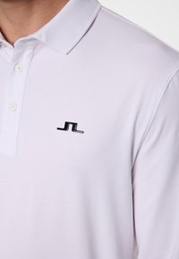 Läheltä kuvattu henkilö, jolla on päällään valkoinen polo-paita, jossa on musta JL-logo vasemmassa rinnassa ja valkoisia nappeja kauluksen läpällä.