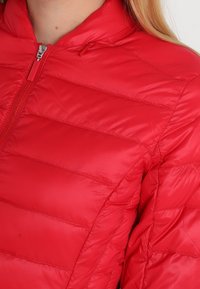 Červená péřová bunda s prošívanou texturou, vysokým límcem a zipovým uzávěrem. Vyrobena z lehkého, vodoodpudivého materiálu.