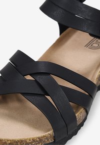Sandale noire avec des sangles synthétiques croisées, semelle en liège et semelle texturée. Présente un design plat et une découpe ouverte au niveau des orteils.
