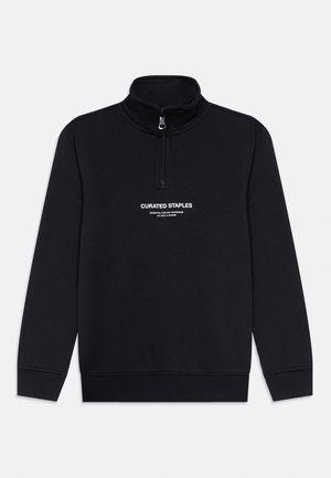 Felpa nera con colletto a metà zip, caratterizzata da testo bianco che recita "CURATED STAPLES" e "ESSENTIAL FOR ANY WARDROBE". Texture morbida e liscia.