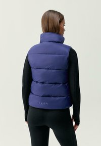 Temno modra pufferska brezrokavnik z visokim ovratnikom, ki vsebuje določenih delovijo in gladko teksturo. Logo je viden blizu roba. Črn oprijet dolgi rokav pod njim.