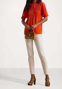 Orange pleated blouse with a tie neck, cream fitted pants, and brown platform sandals. The model holds a small woven bag with a clasp.