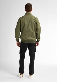 Jachetă bomber verde măsliniu cu o croială dreaptă și manșete ribbrate, asociată cu pantaloni cargo negri cu buzunare la genunchi și încălțăminte bej.