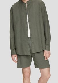 Man in een olijfgroene linnen overhemden met knopen en bijpassende shorts, met een wit onderhemd, staande tegen een effen achtergrond.