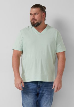 Mann mit Bart und Haarknoten, der ein hellgrünes V-Ausschnitt-T-Shirt und blaue Jeans trägt, vor einem schlichten hellgrauen Hintergrund stehend.