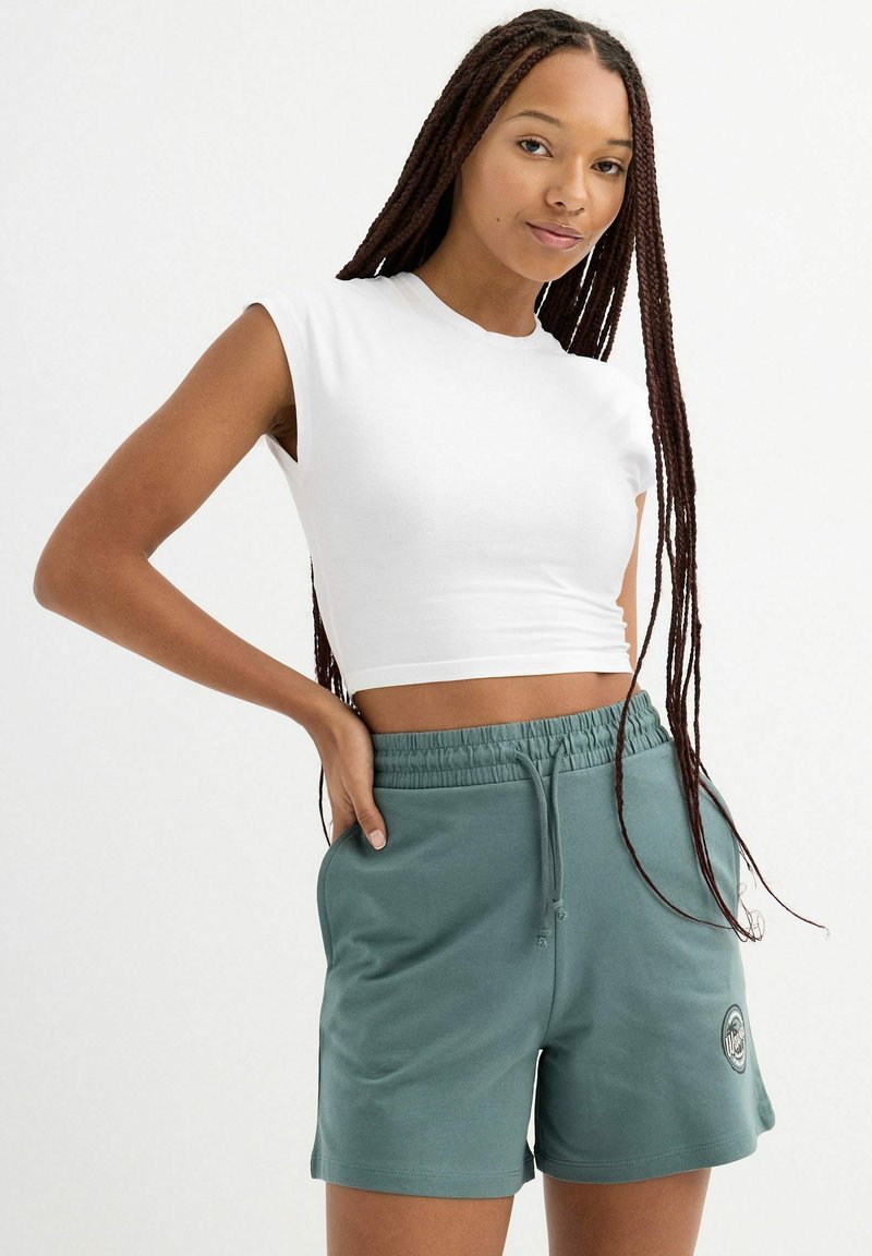 Top sin mangas blanco cortado combinado con pantalones cortos de tiro alto en teal con cordón, que incluyen un parche con logo y bolsillos laterales, fabricados en un tejido suave.