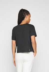 Moteris su pintomis plaukais, vilkinti juodą trumpą rankovę viršutinį drabužį su šoninėmis išpjovomis ir baltas kelnes, stovi su viena ranka kišenėje, atsisukusi atgal.