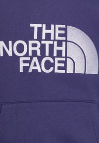 Sudadera púrpura con gran logo en blanco "THE NORTH FACE" y diseño en semicírculo, fabricada en tejido suave y texturizado con un bolsillo delantero.