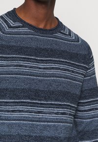 Námořnický modrý pletený svetr s horizontálními pruhy v různých odstínech modré a bílé. Má ribbovaný kulatý výstřih a texturovanou tkaninu.