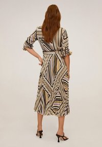 Femme aux longs cheveux rouges portant une robe mi-longue à rayures géométriques en beige, noir et lavande, accompagnée de sandales à talons noires.