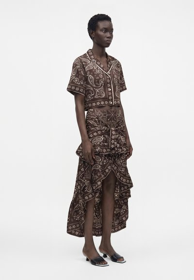 Mannequin portant une chemise courte marron à manches courtes avec motif cachemire et une jupe volantée asymétrique assortie, avec des talons noirs ouverts à l'avant et bas.