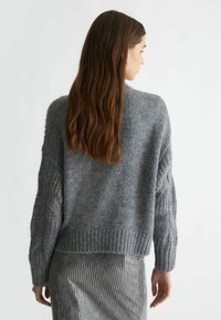 Maglione grigio e morbido con orlo a costine e maniche testurizzate, indossato sopra una gonna argento scintillante, visto da dietro.