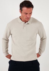 Polo din bumbac bej, cu mâneci lungi, textură aleasă, guler clasic, plachetă cu două butoane și manșete în contrast, plasat pe un fundal simplu.