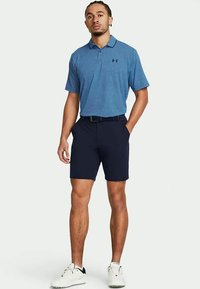 Uomo che indossa una polo blu a pois, pantaloni corti neri con cintura e scarpe da ginnastica bianche, in piedi con le mani nelle tasche.