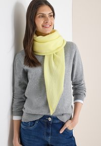 Écharpe jaune clair avec un motif texturé, portée lâchement autour du cou sur un pull gris. L'écharpe présente une forme triangulaire et un tissu doux.