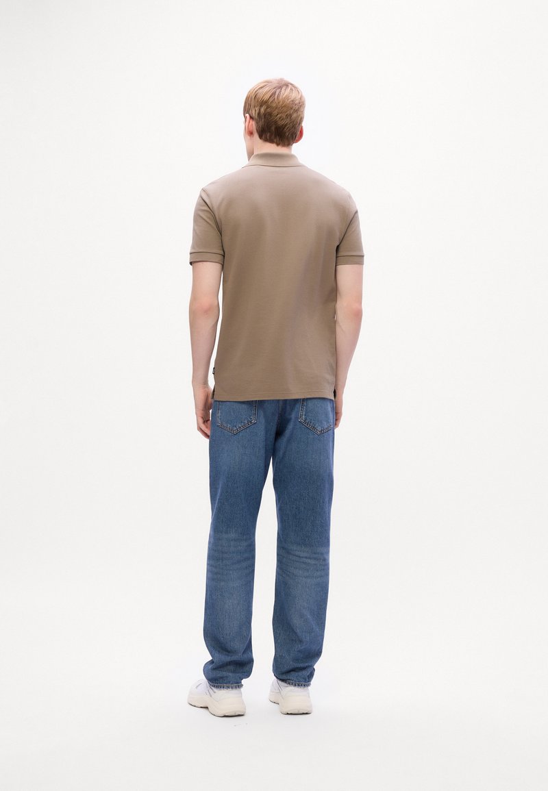 Homme debout de dos portant un polo marron à manches courtes, un jean bleu et des baskets blanches sur un fond blanc.