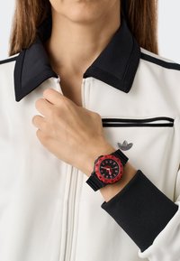 Must musta ja punane kell ümmarguse näo, julged tunni märgid ja kummirihmaga, kantud käel, mis toetub valge ja musta tõmblukuga jope vastu.