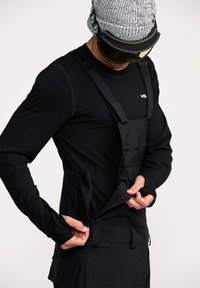 Zwarte sneeuwoverall met banden, gemaakt van duurzaam materiaal. Beschikt over meerdere zakken en een ritssluiting. Model draagt een gebreide beanie en een skibril.