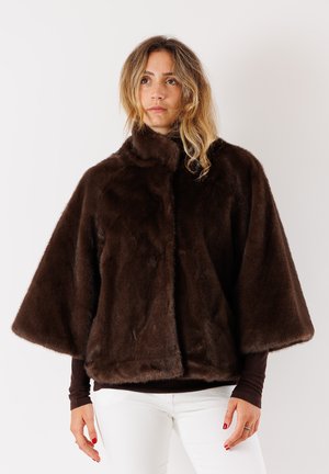 Cappotto in pelliccia sintetica marrone con un alto colletto, maniche larghe e una texture morbida, posizionato sopra un top scuro e pantaloni bianchi.