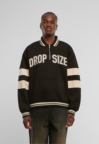 Zwarte zip-up sweatshirt met "DROP SIZE" in vette witte letters gedrukt. Voorzien van gestreepte manchetten en onderrand, en een voorzak. Textuur materiaal.