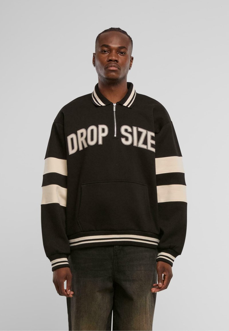 Zwarte zip-up sweatshirt met "DROP SIZE" in vette witte letters gedrukt. Voorzien van gestreepte manchetten en onderrand, en een voorzak. Textuur materiaal.