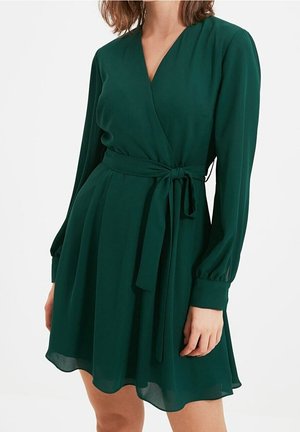 Mujer con un vestido cruzado de manga larga de color verde oscuro con un cinturón de tela a juego atado a la cintura, de pie frente a un fondo liso.